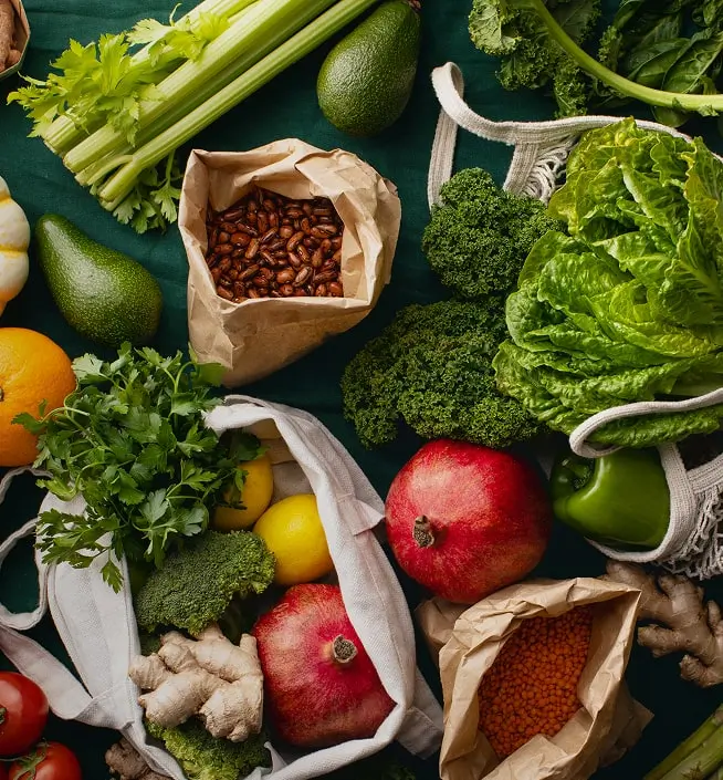Assorted fresh vegetables, fruits, beans, and lentils arranged on a green surface, with some items in reusable bags and paper bags.