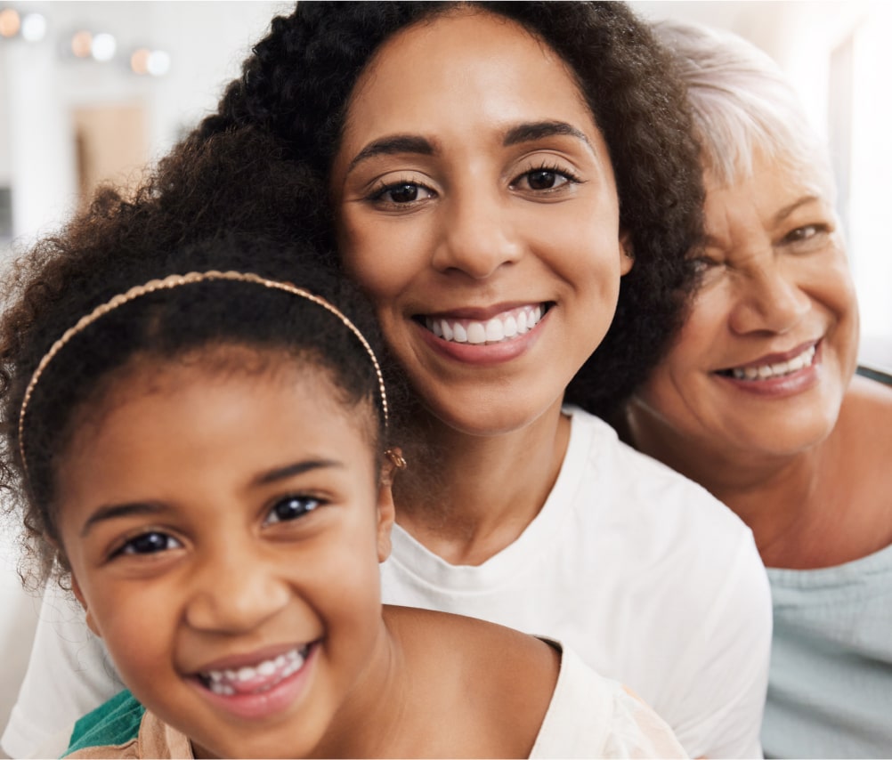 Three generations of women—child, adult, and senior—smiling close together indoors, facing the camera.