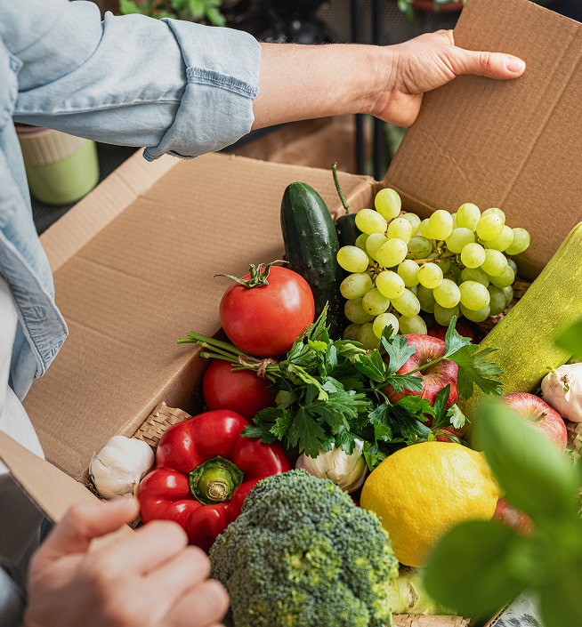 A person holds an open cardboard box filled with assorted fresh fruits and vegetables, including tomatoes, grapes, broccoli, and a lemon.