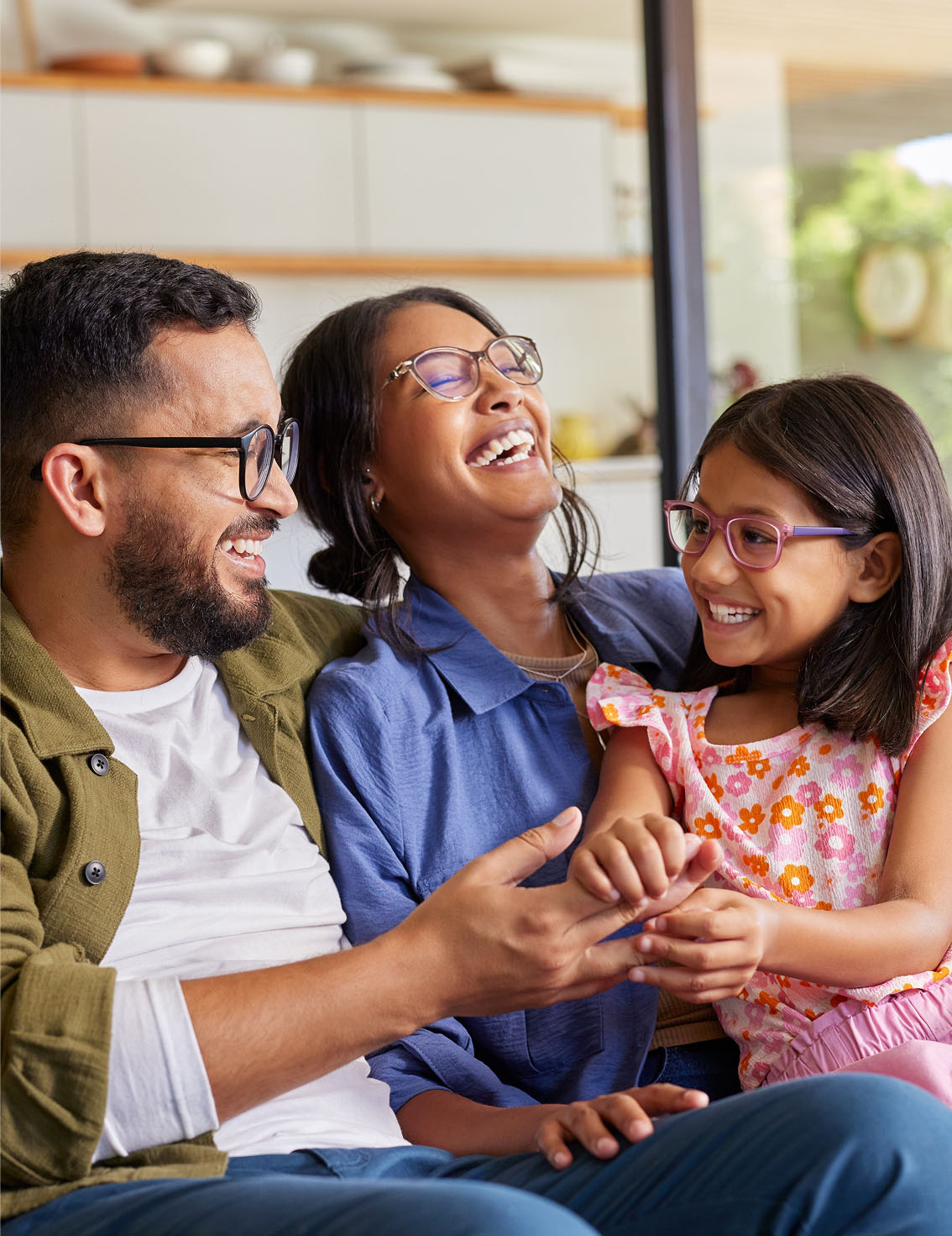 A man, woman, and young girl sit close together on a couch, smiling and laughing in a bright, modern living room.