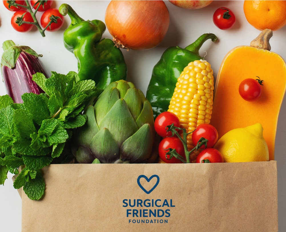A paper bag labeled "Surgical Friends Foundation" is filled with various fresh vegetables and fruits, including corn, tomatoes, mint, peppers, lemon, and squash.