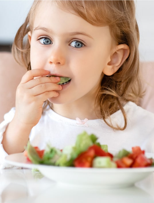 A young child with light brown hair and blue eyes eats salad with her hand, sitting at a table with a plate of vegetables in front of her.