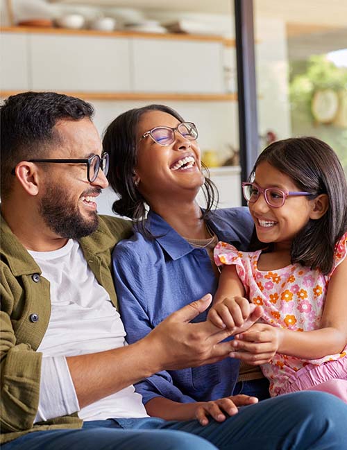 Three people, two adults and a child, sit closely together on a couch indoors, smiling and laughing.